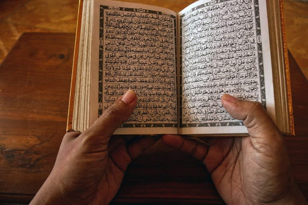 Deux mains tiennent un Coran ouvert, aux pages couvertes d'écriture arabe, au-dessus d'une table en bois