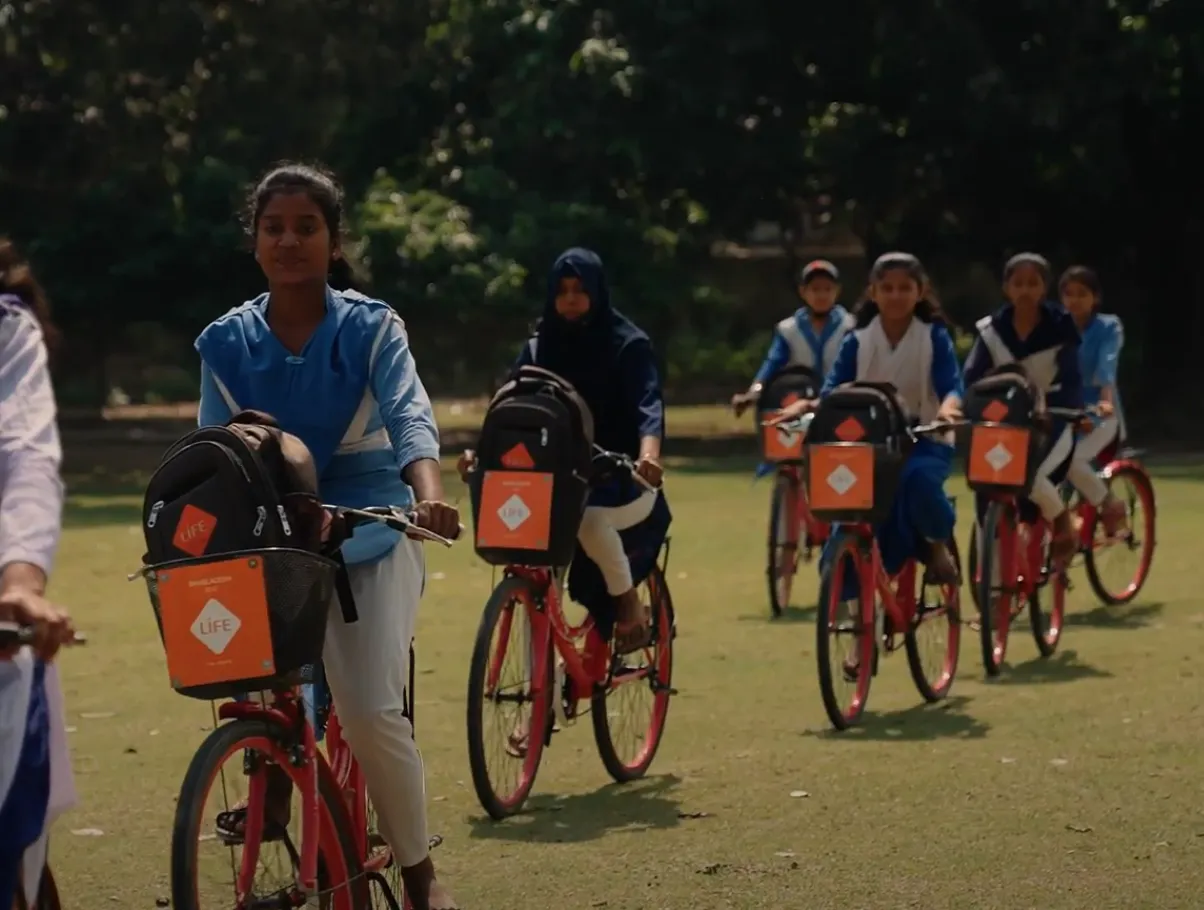 Des écolières à vélo rouge, avec sac à dos et panier orange portant le logo LIFE, roulent sur une pelouse