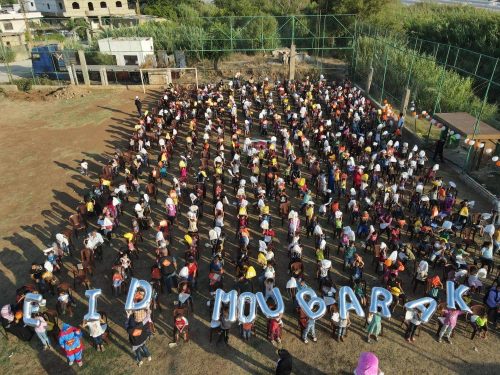 Vue aérienne d'une foule d'enfants assis en rangs sur un terrain, avec des ballons formant les mots « EID MOUBARAK »