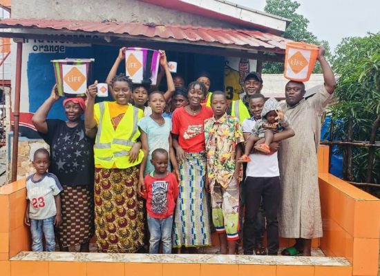 Groupe d'adultes et d'enfants souriants devant une maison, brandissant des seaux et tasses LIFE sur un muret orange