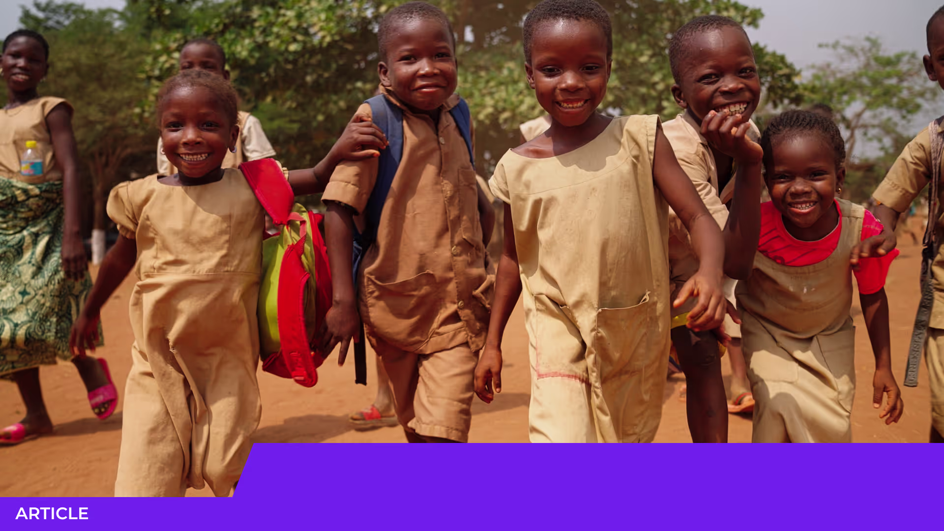 Enfants souriants en uniforme beige marchant ensemble sur un chemin de terre, l'un portant un cartable rouge