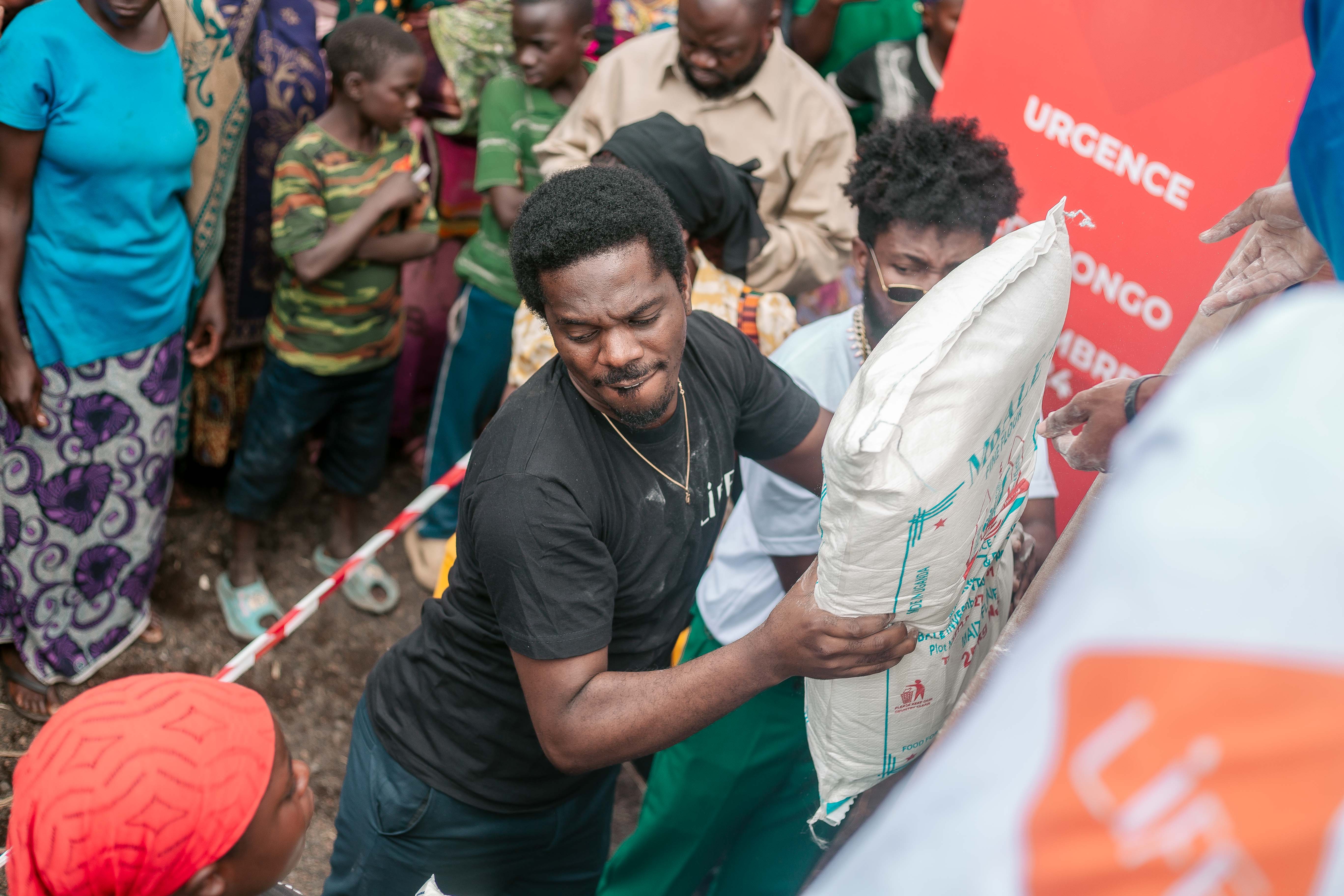 Un bénévole en t-shirt LIFE porte un sac de riz lors d'une distribution d'urgence, entouré d'une foule d'adultes et d'enfants