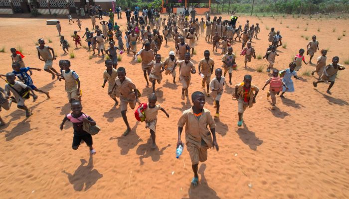 Des dizaines d'enfants en uniforme beige courent sur une cour de terre rouge, vus d'en haut, devant une école