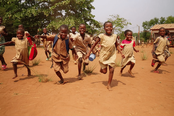 Enfants en uniforme beige courant et riant dans une cour de terre rouge d'une école, sacs sur le dos, arbres derrière eux