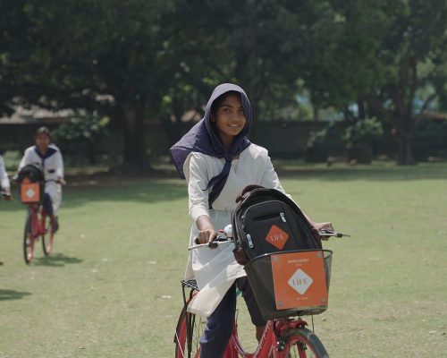 Jeune fille souriante au foulard sur un vélo rouge, sac à dos dans le panier orange portant le logo LIFE, une autre cycliste derrière
