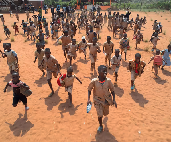 Vue du dessus d'une foule d'enfants en uniforme beige courant sur une cour de terre rouge, sous le soleil