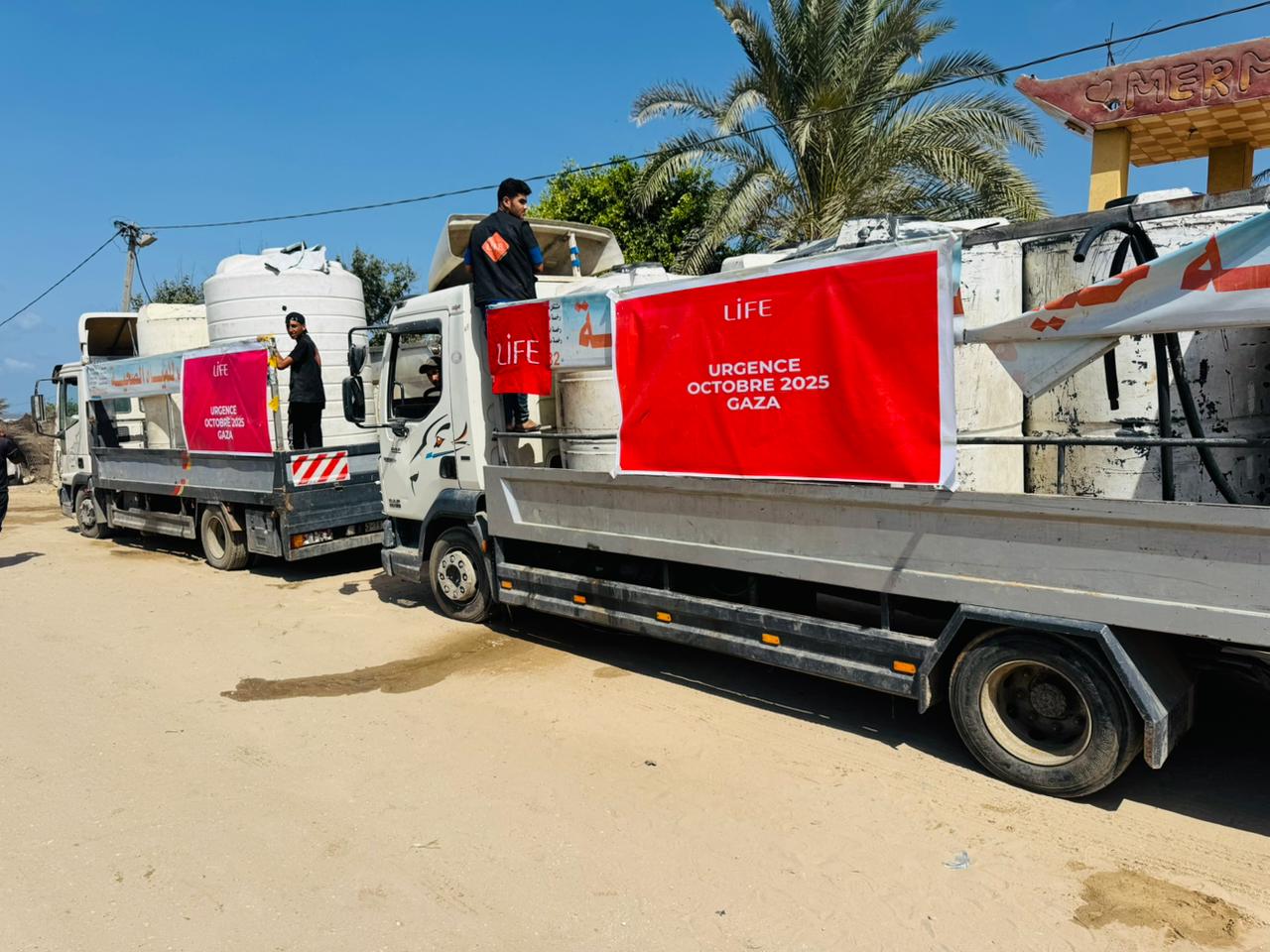 Deux camions-citernes avec banderoles rouges LIFE « Urgence octobre 2025 Gaza » garés sur un chemin de sable sous un ciel bleu.