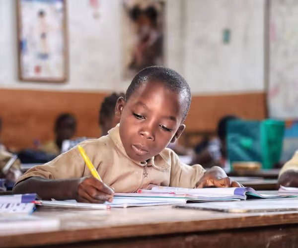 Un jeune garçon en uniforme beige écrit avec un crayon jaune sur un cahier, assis à son pupitre dans une salle de classe