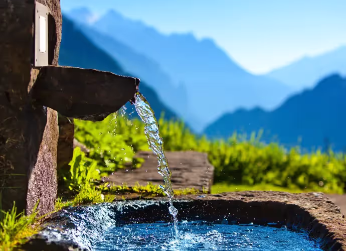 Fontaine en pierre où l'eau s'écoule dans un bassin, avec des montagnes bleutées et de l'herbe verte au fond