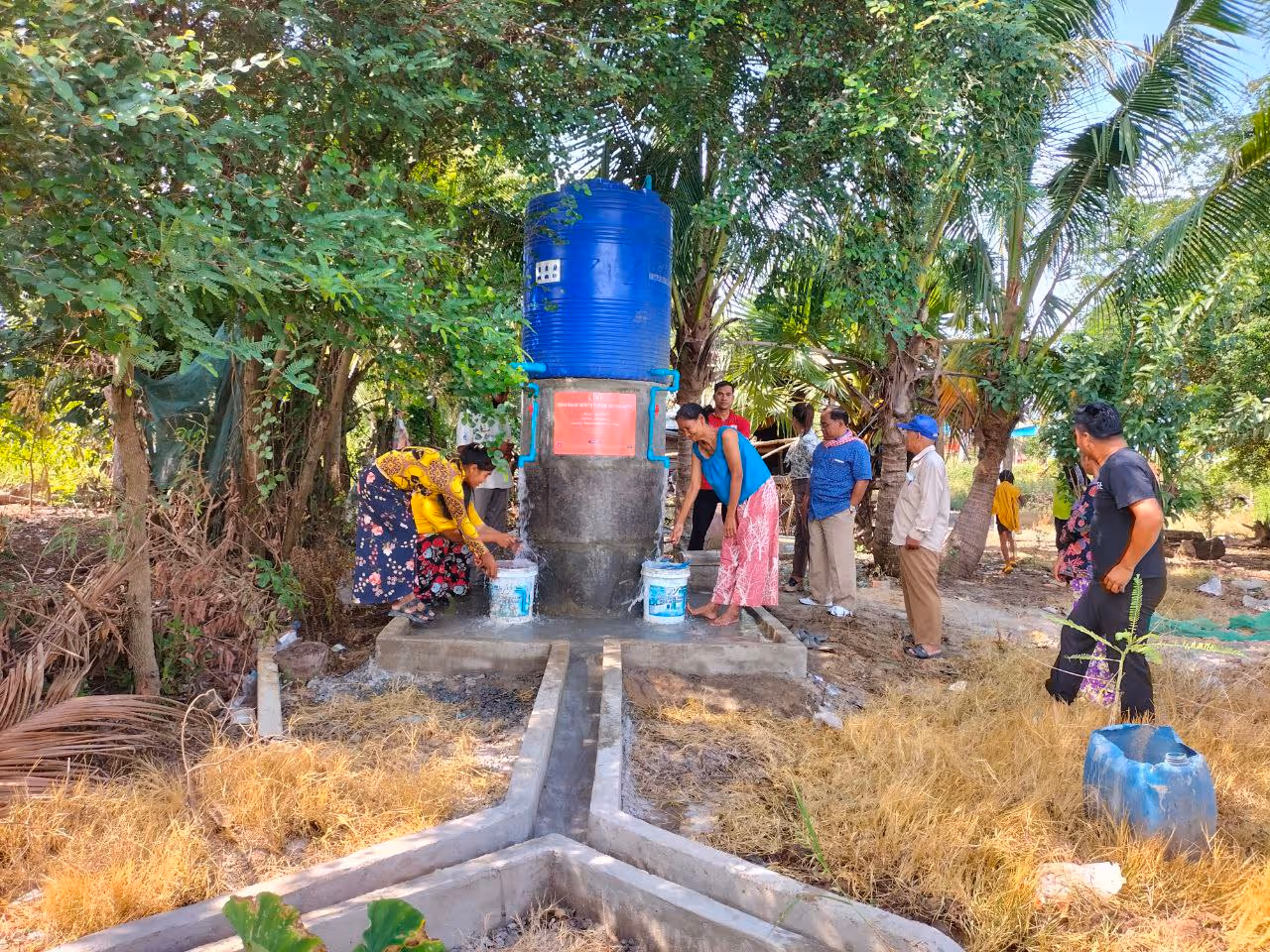 Habitants remplissant des seaux à un point d'eau en béton surmonté d'un grand réservoir bleu, dans un jardin ombragé de palmiers