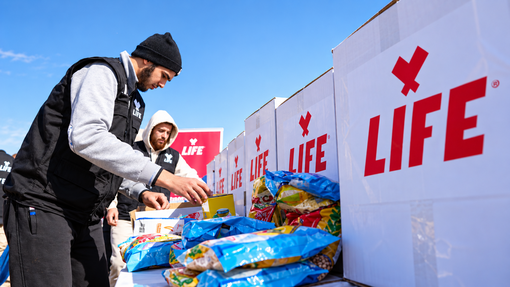 Deux bénévoles LIFE en gilet noir préparent des denrées près de grands cartons blancs portant le logo LIFE
