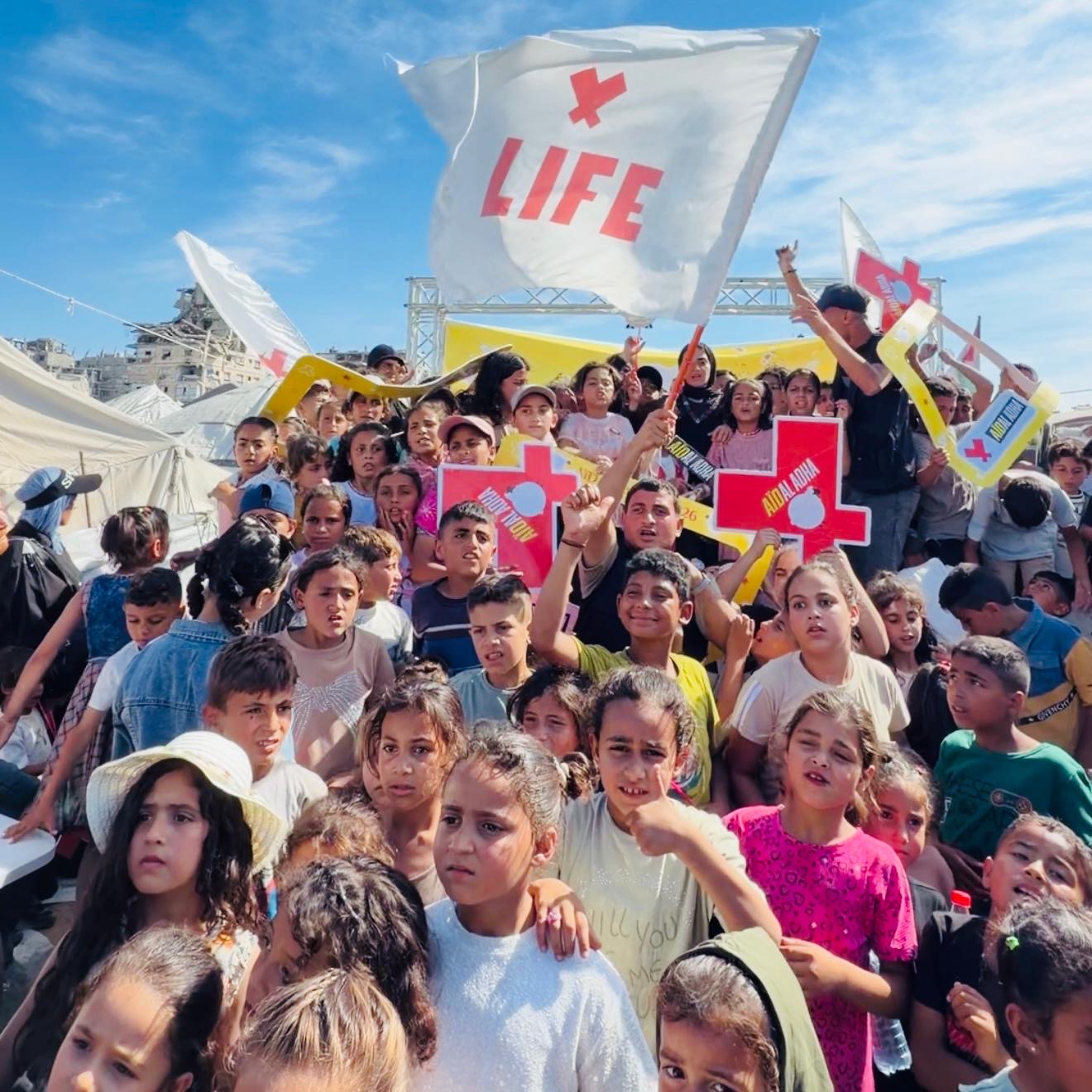 Foule d'enfants rassemblés sous un grand drapeau blanc LIFE, avec des panneaux en forme de croix rouge, près de tentes