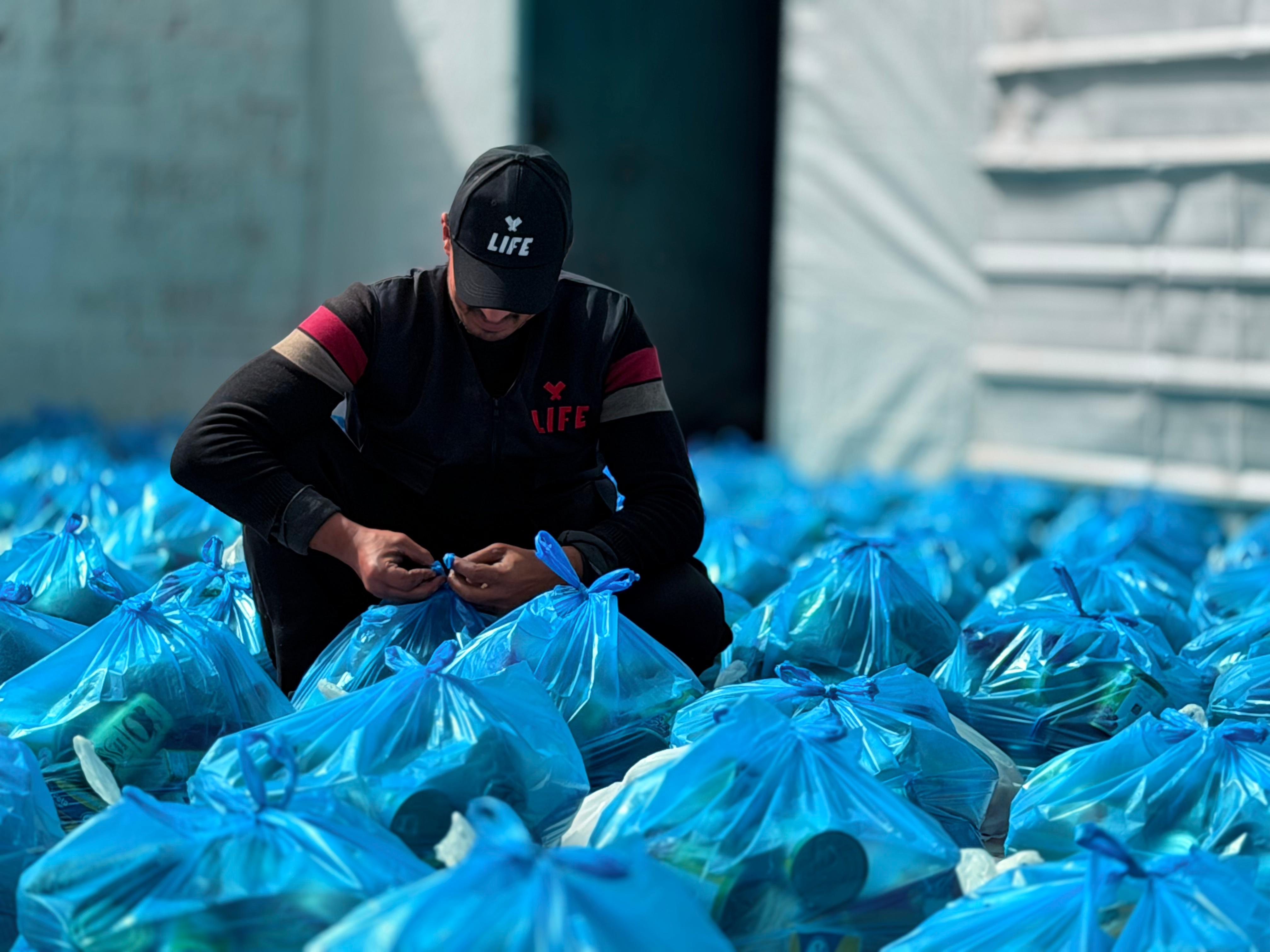Un bénévole LIFE casquette noire noue des sacs bleus remplis de denrées, posés en grand nombre au sol