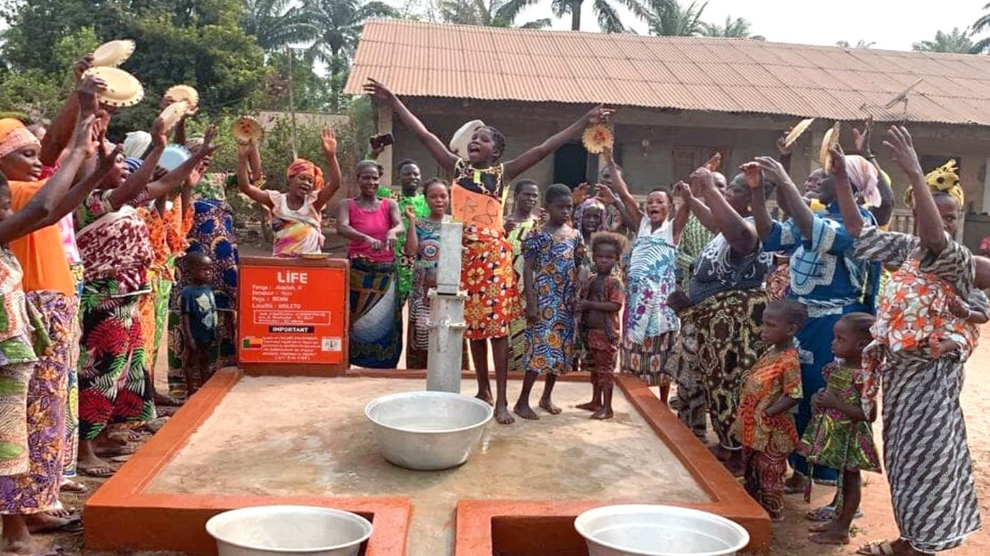 Villageois en tenue colorée, bras levés et assiettes en main, célébrant autour d'une pompe à eau LIFE dans un village