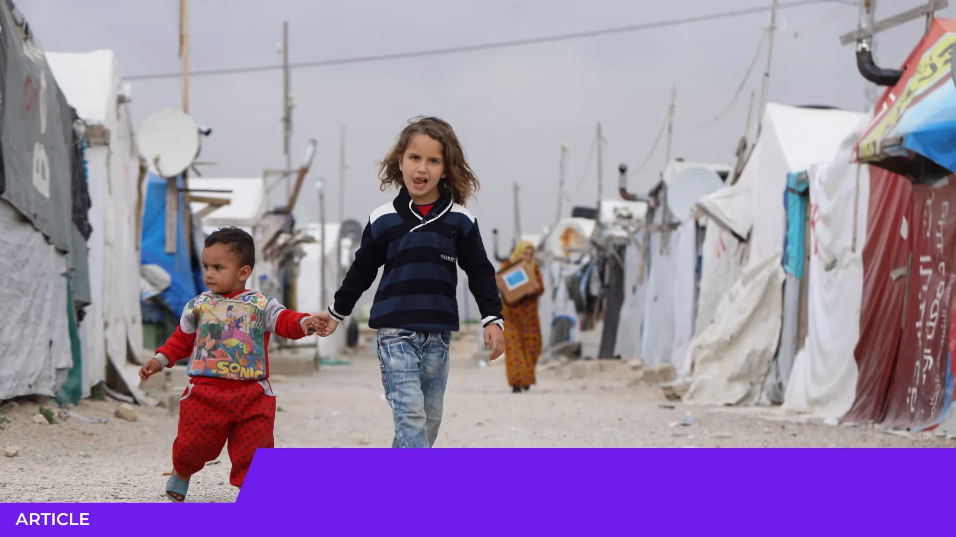 Deux enfants se tiennent par la main et marchent dans une allée entre des tentes d'un camp de réfugiés, sous un ciel gris
