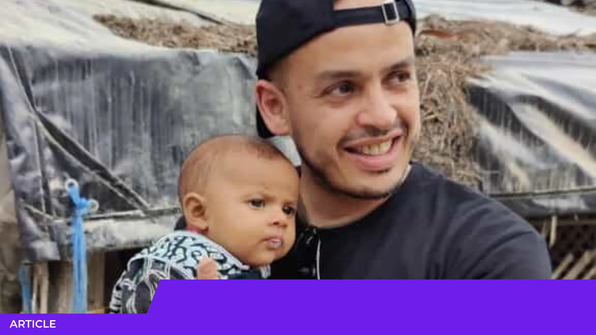 Un homme souriant en casquette noire tient un jeune bébé dans ses bras devant des bâches et de la paille