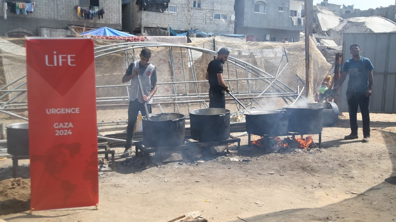 Des hommes remuent de grandes marmites sur des feux, à côté d'une bannière LIFE « Urgence Gaza 2024 »