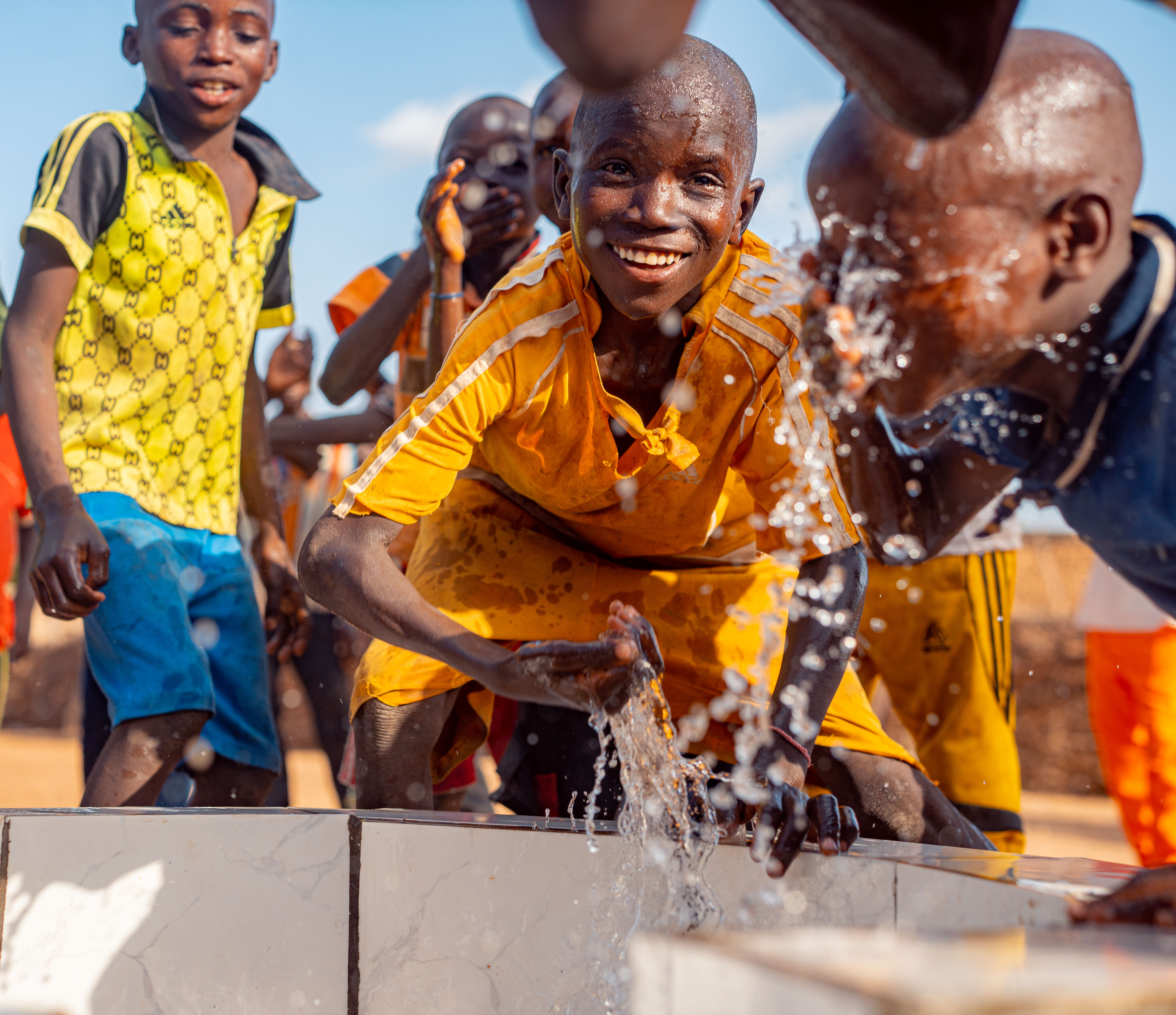 Enfants souriants s'aspergeant d'eau qui jaillit d'un point d'eau, en maillots jaunes, sous un ciel bleu