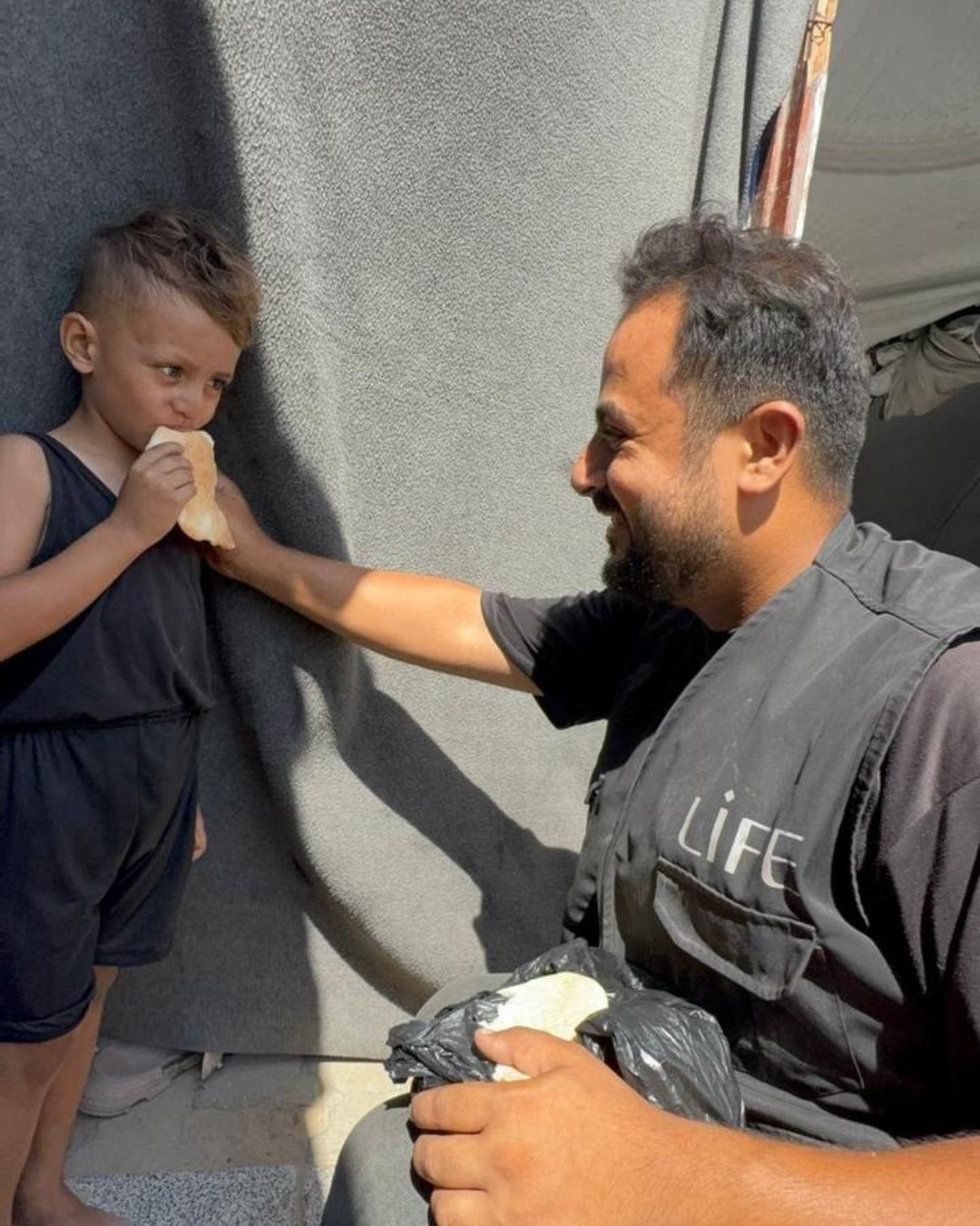 Un bénévole LIFE sourit à un jeune enfant qui mange un morceau de pain, devant une paroi de tente grise