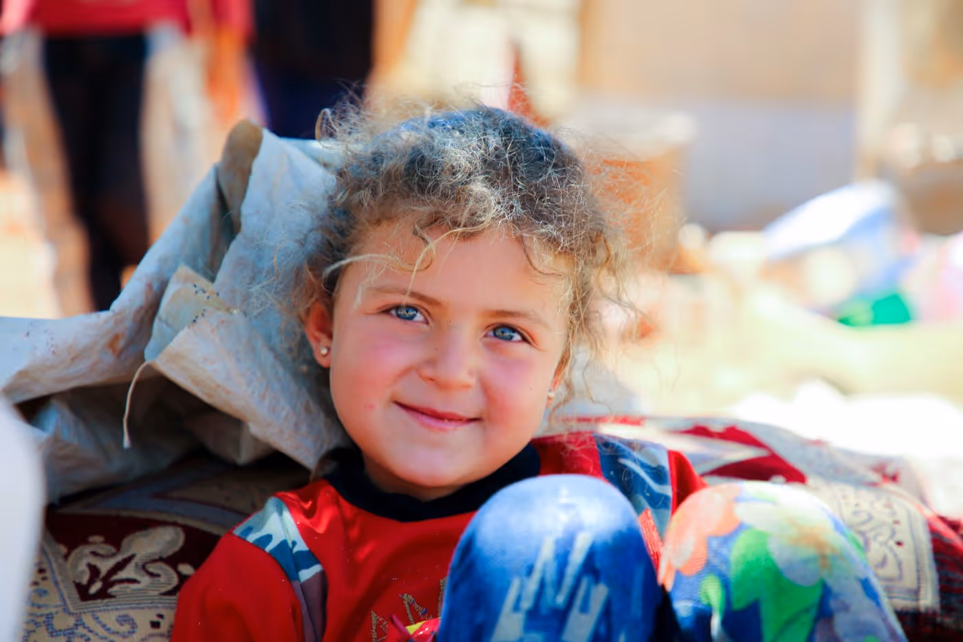 Fillette aux cheveux bouclés et aux yeux clairs, souriante, assise sur des tapis et des sacs, vêtue d'un haut rouge