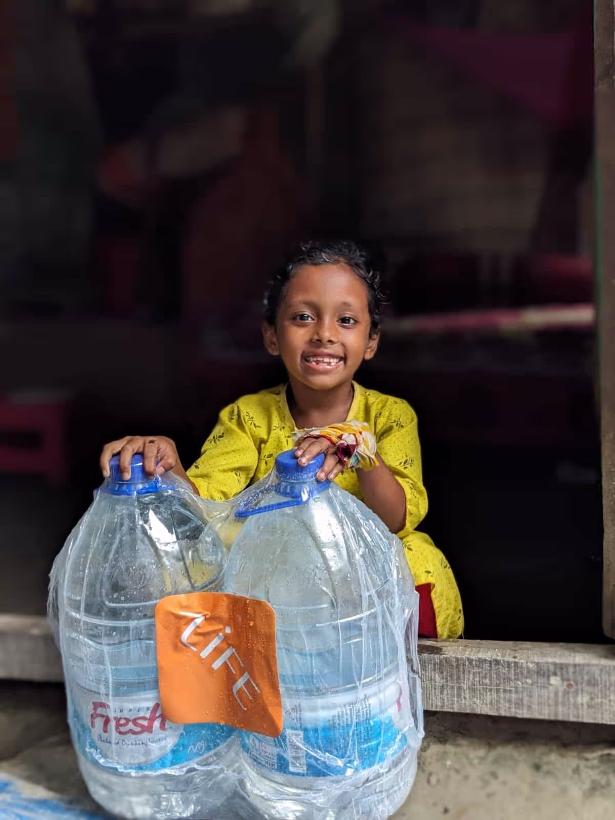 Petite fille souriante en robe jaune, assise sur un rebord, les mains posées sur deux grands bidons d'eau potable portant l'étiquette LIFE.