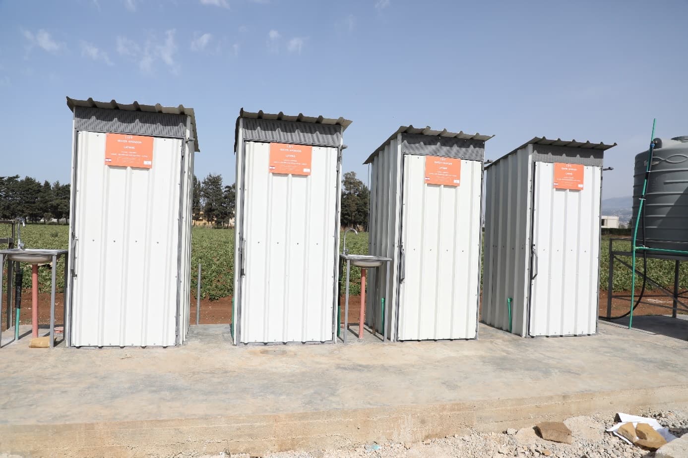 Quatre latrines en tôle blanche alignées, chacune avec un panneau orange LIFE, et des lavabos, au bord d'un champ