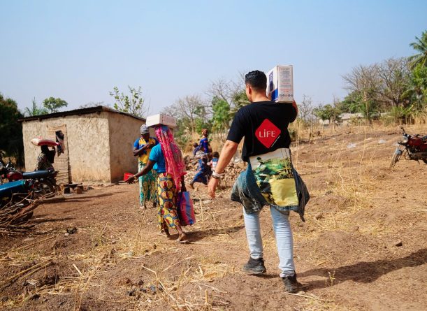 Un volontaire au t-shirt LIFE, de dos, porte un colis sur l'épaule sur un chemin de terre, près de femmes chargées de colis