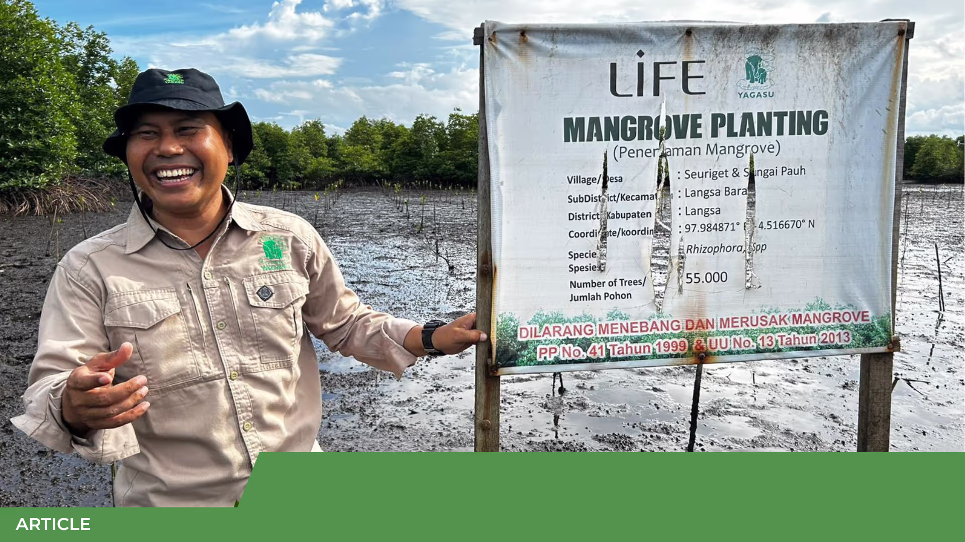 Homme souriant en tenue beige devant un panneau LIFE « Mangrove Planting » dans une mangrove boueuse aux jeunes plants