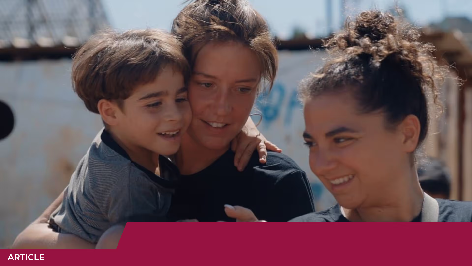 Un petit garçon porté dans les bras d'une femme souriante, à côté d'une autre femme qui sourit, en extérieur
