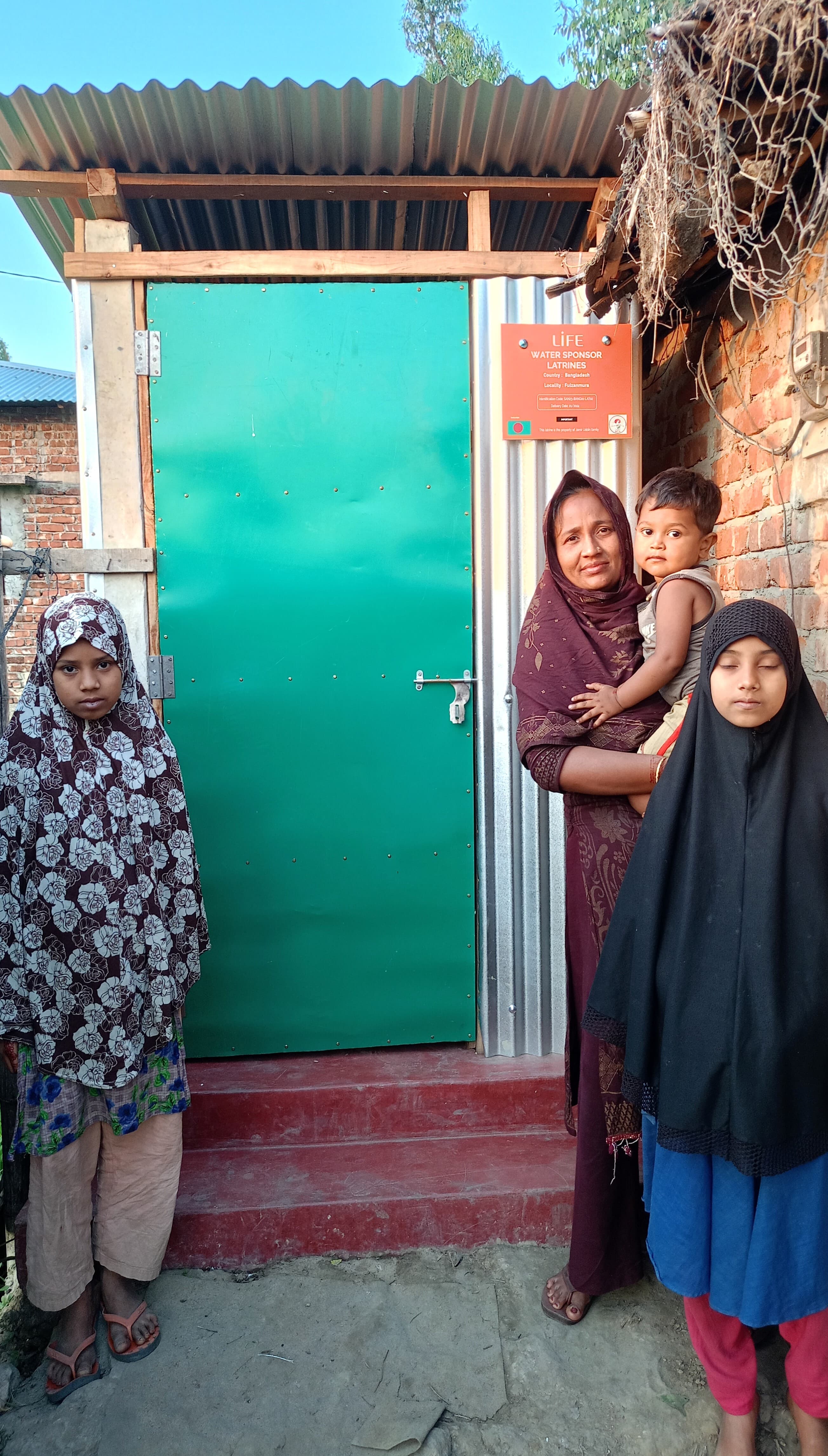 Une femme portant un enfant et deux jeunes filles posent devant une latrine à porte verte, avec un panneau LIFE