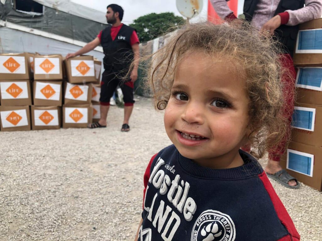 Un jeune enfant aux cheveux bouclés sourit à la caméra devant des cartons LIFE empilés, dans un camp au sol de gravier.