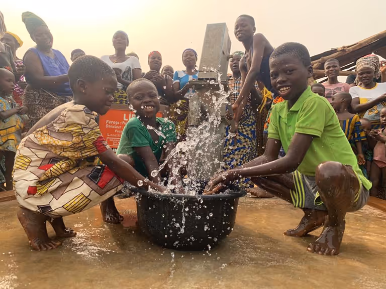 Deux enfants accroupis éclaboussent l'eau d'une bassine sous une pompe, entourés d'une foule de villageois souriants.