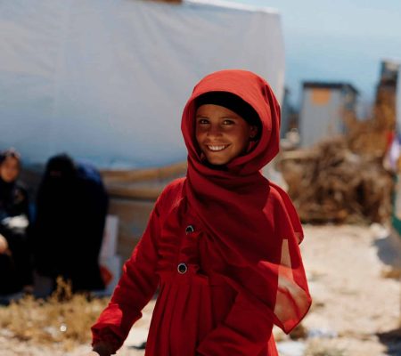 Jeune fille souriante vêtue d'un manteau et d'un voile rouges, devant une tente blanche dans un camp