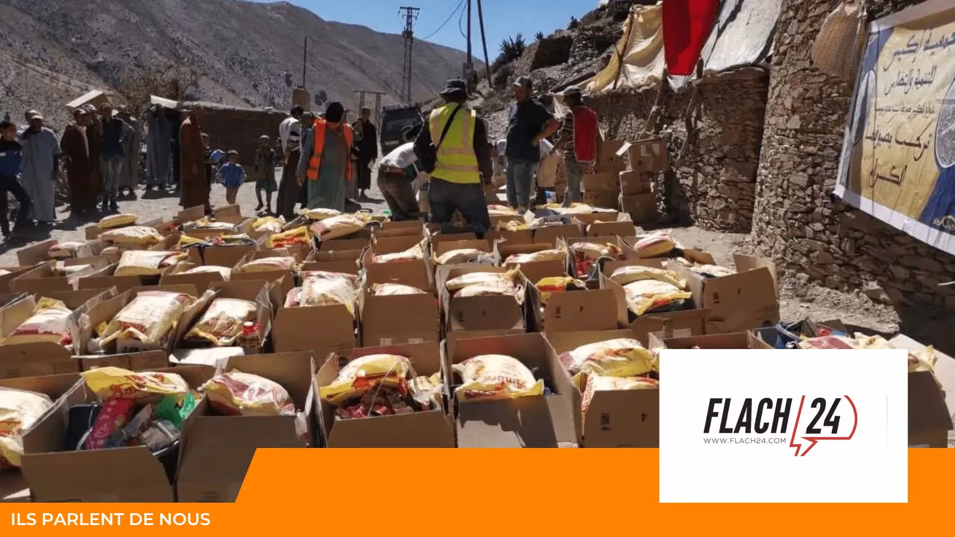 Distribution de colis alimentaires en cartons alignés dans un village de montagne, avec des bénévoles en gilets fluo