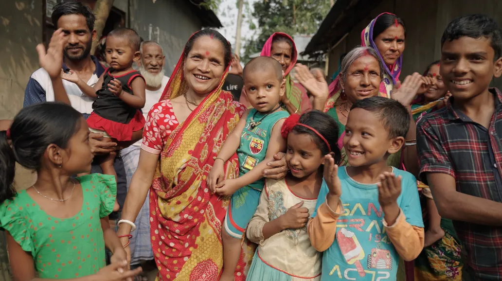 Groupe d'enfants, de femmes et d'hommes souriant et applaudissant devant des habitations, une femme en sari rouge au centre