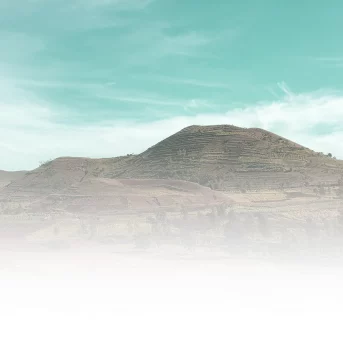 Colline aux flancs aménagés en terrasses, sur un sol aride, sous un ciel turquoise voilé de nuages et de brume
