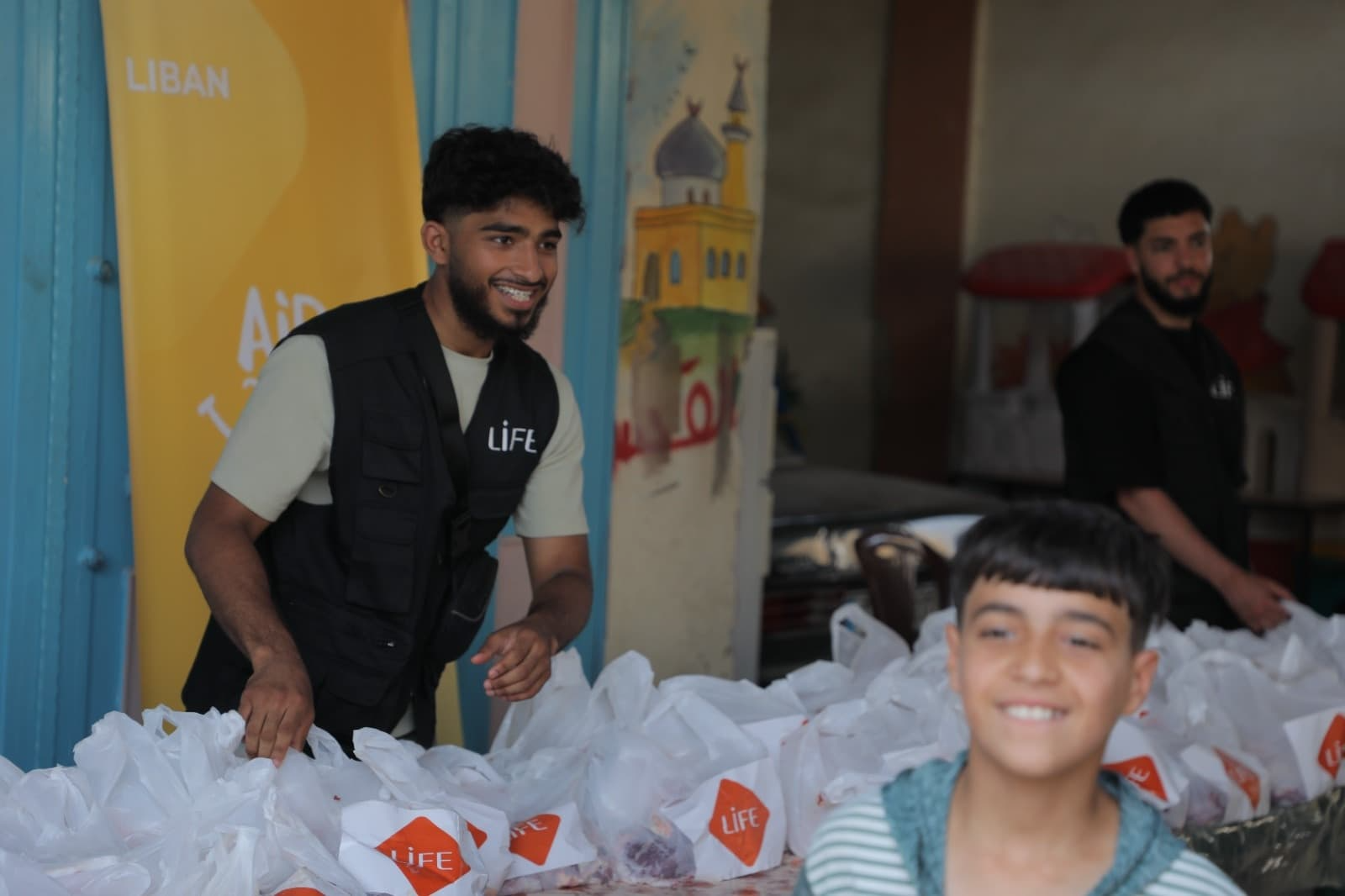 Un bénévole LIFE en gilet noir sourit derrière une table de sacs blancs de distribution, avec un garçon souriant au premier plan