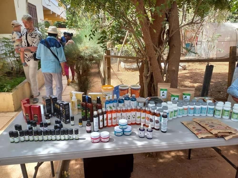 Table dans un jardin avec de nombreux produits : huiles essentielles, flacons, crèmes, pots et sachets ; des visiteurs au fond.