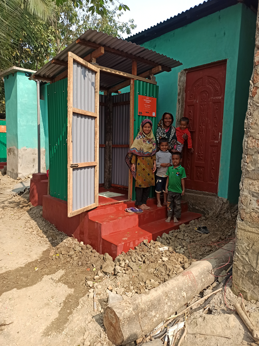 Une femme et trois enfants posent devant une latrine neuve en tôle, aux marches rouges, avec un panneau LIFE