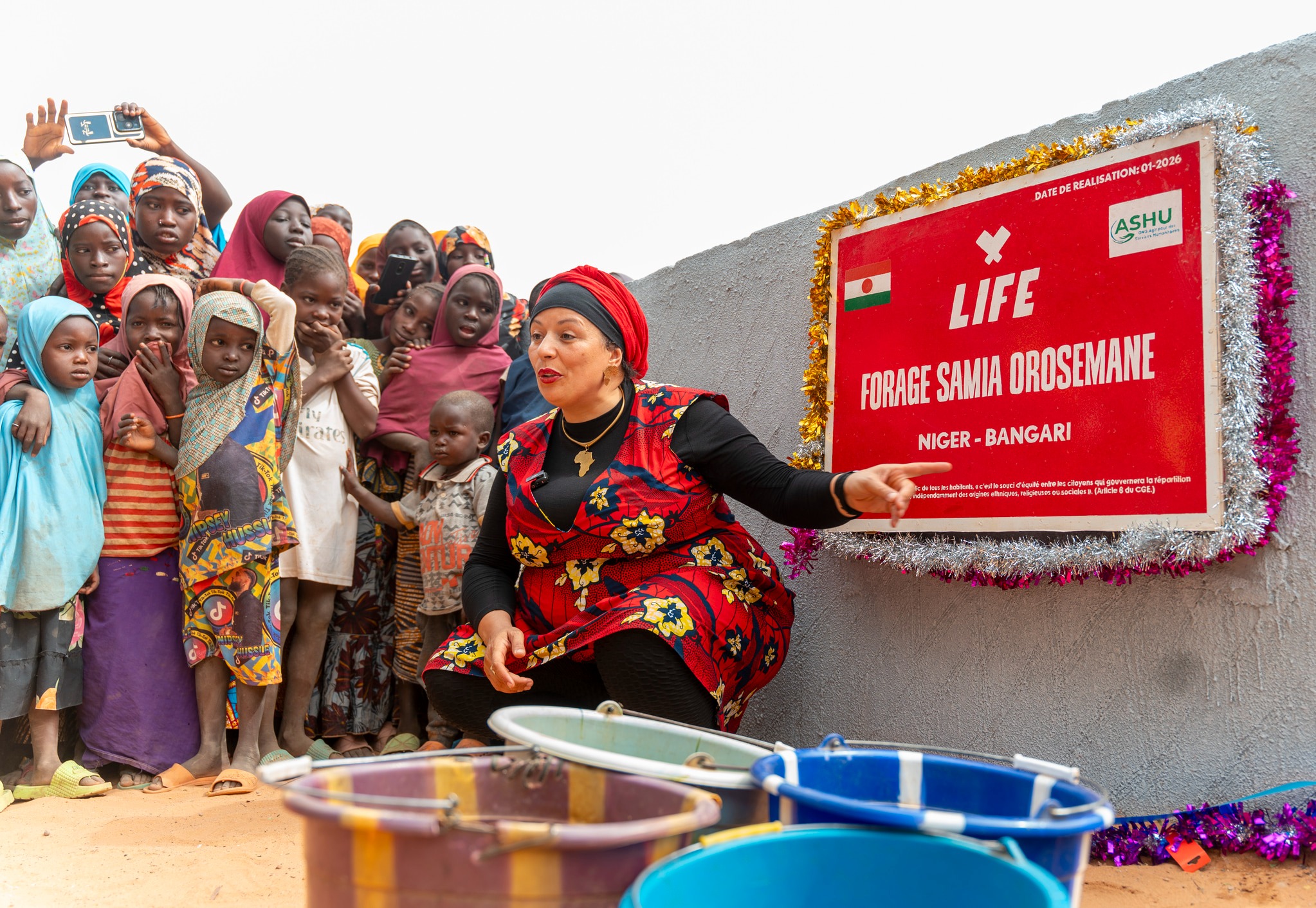 Femme accroupie devant un panneau LIFE « Forage Samia Orosemane, Niger - Bangari », entourée d'enfants et de seaux