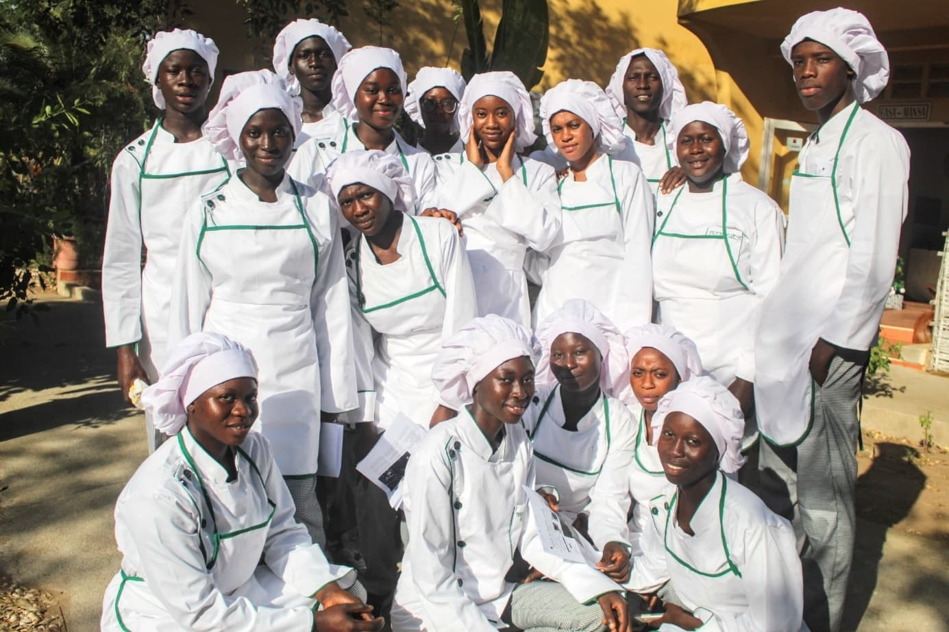Groupe de jeunes en tenue de cuisinier blanche à liserés verts et toque, posant ensemble en extérieur, certains accroupis