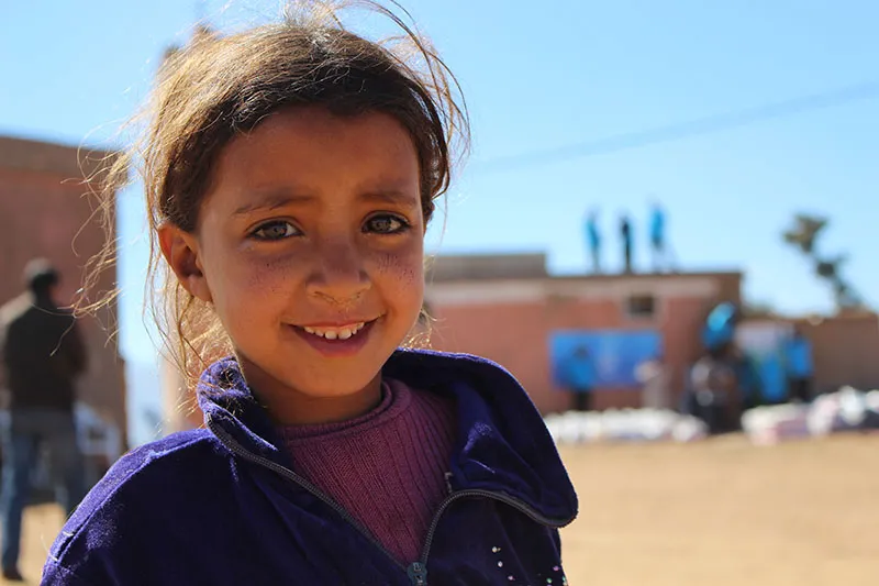 Portrait souriant d'une petite fille en veste violette, cheveux au vent, devant des bâtiments où des personnes en bleu travaillent