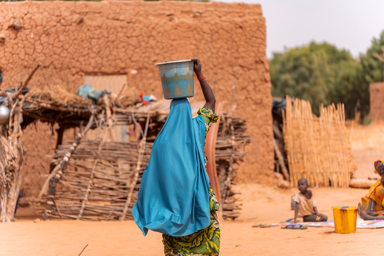 Femme de dos portant un seau sur la tête dans un village de maisons en terre, un enfant assis au sol à droite