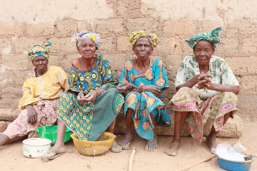 Quatre femmes âgées en tenues colorées assises côte à côte devant un mur en terre, avec des bassines et un panier devant elles