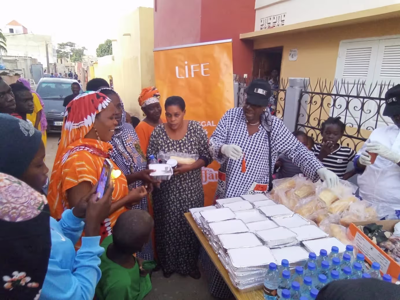 Distribution de repas en barquettes et de bouteilles d'eau dans une rue, devant une bannière LIFE, avec des femmes et des bénévoles gantés