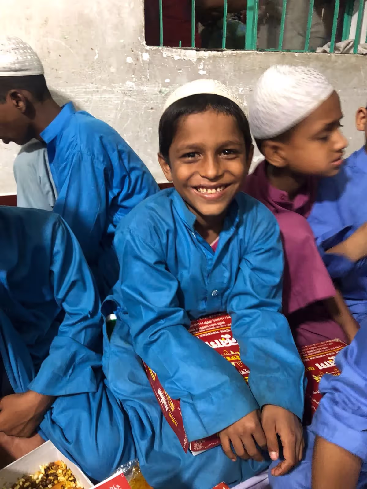 Un garçon souriant en tenue bleue et calotte blanche, assis parmi d'autres enfants, tenant des paquets de nourriture
