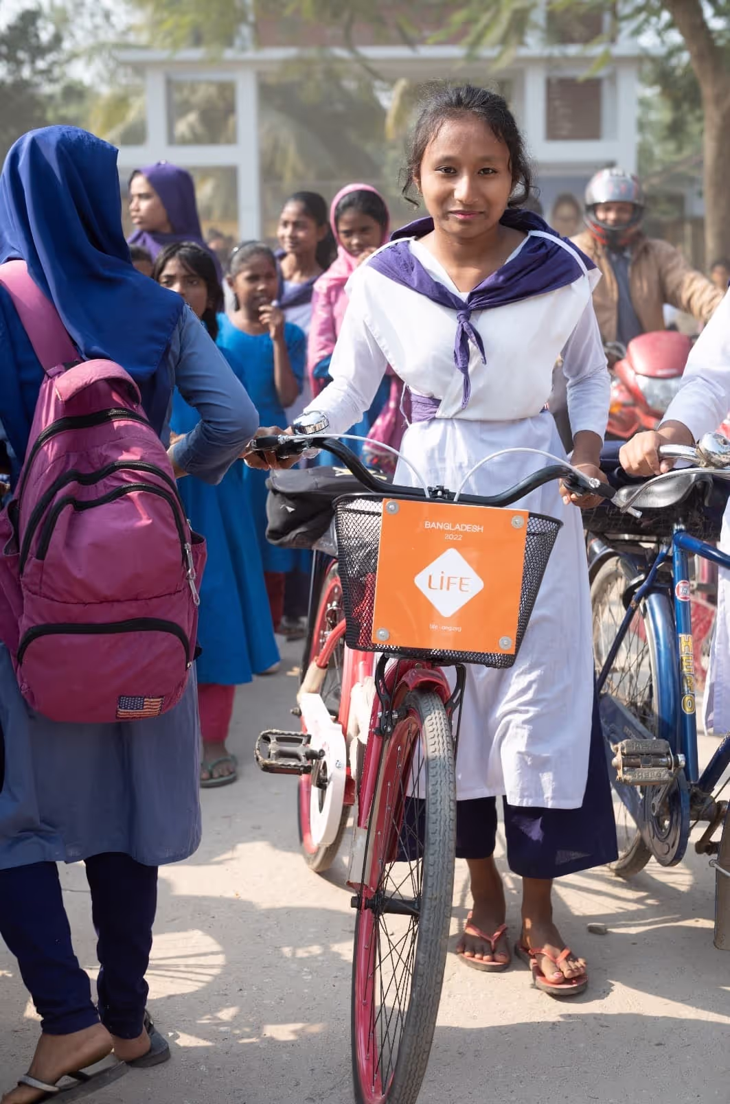 Une écolière en uniforme blanc et violet tient un vélo dont le panier porte une plaque LIFE, entourée d'autres élèves dans la rue.