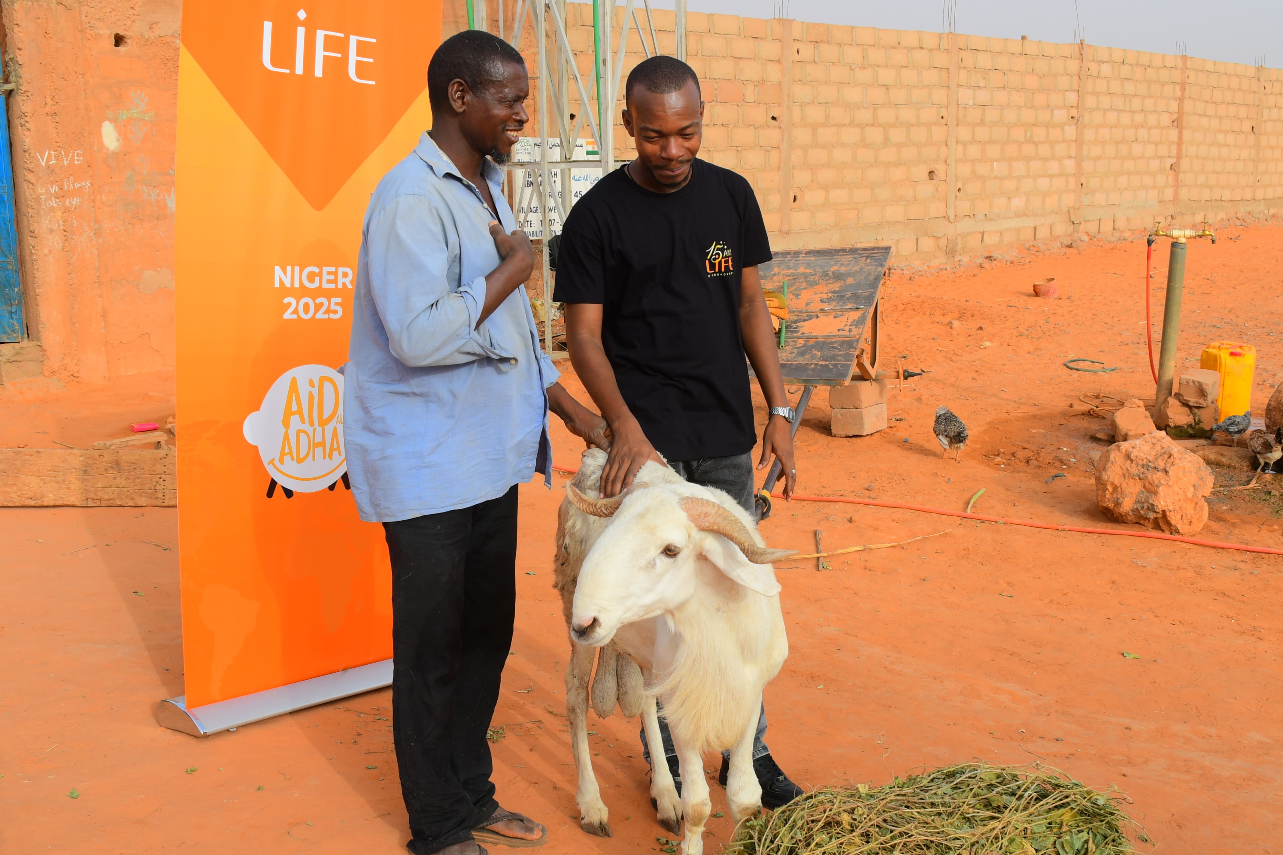Deux hommes souriants caressent un mouton blanc devant une bannière LIFE « Niger 2025 – Aïd Adha », sur un sol de terre rouge