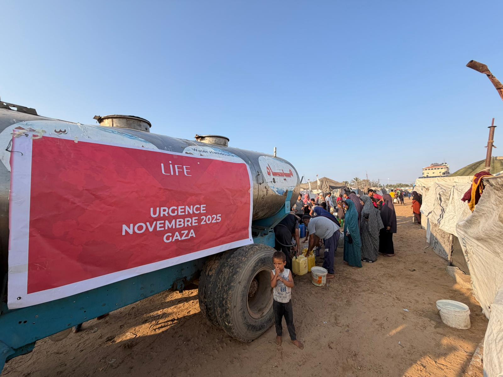 Distribution d'eau par un camion-citerne portant une bannière LIFE « Urgence novembre 2025 Gaza », devant une foule
