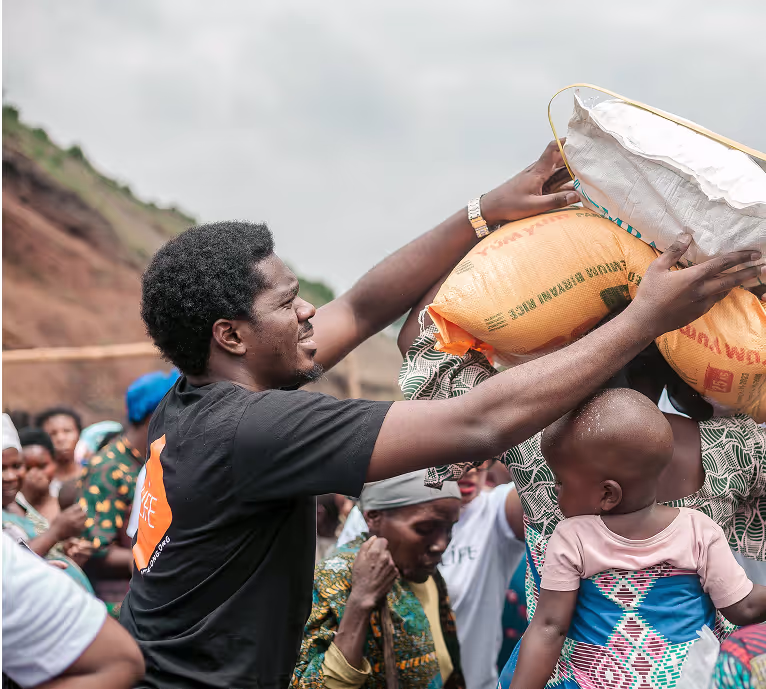 Un membre de LIFE en t-shirt noir aide une femme à porter un sac de riz sur la tête lors d'une distribution, avec un enfant sur le dos
